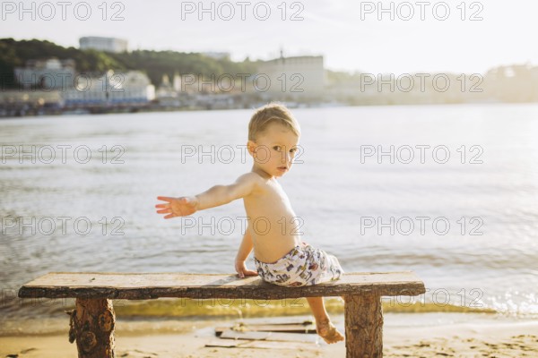 Boy child sitting on a wooden bench with his back on the beach near the water and showing his hand forward direction against the backdrop of the city of Kiev