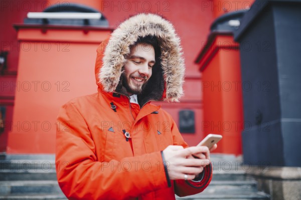Handsome young male student with toothy smile and beard stands on red wall background, facade of educational institution in red winter jacket with hood with fur, Uses finger on screen of mobile phone