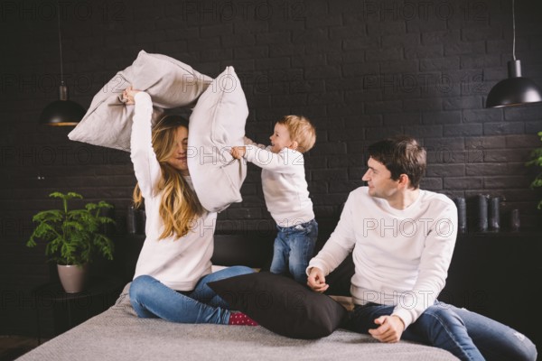 Parents with smiling child playing pillow fight on bed, happy family having fun in bedroom, enjoying leisure activity, funny weekend lifestyle. Happy family having fun in bedroom, pillow battle