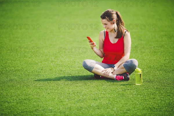 Theme sport health. beautiful young girl sitting resting on green grass lawn stadium using technodogies. In handphone in ear headphones listens music, summer sportswear smart watch heart rate monitor