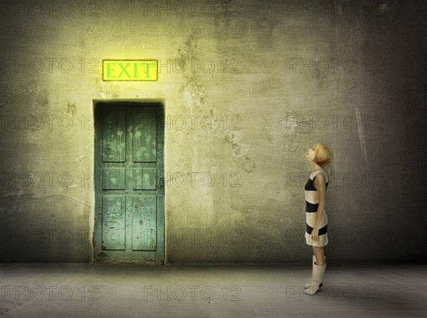 Young woman in black and white dress stand in front of blue door in dark grungy room watch on glowing exit sign, find the way out