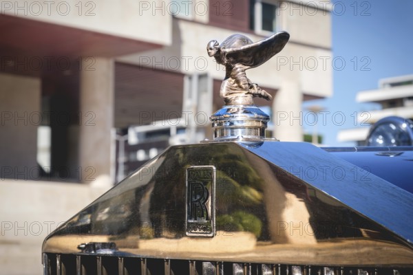 Lisbon, Portugal - Sep 29, 2024: Close-up and selective focus on the Rolls-Royce mascot hood ornament or bonnet badge called Spirit of Ecstasy, female in billowing cloth like wings behind, leaning forward, and emblem on grille. Rare kneeling version