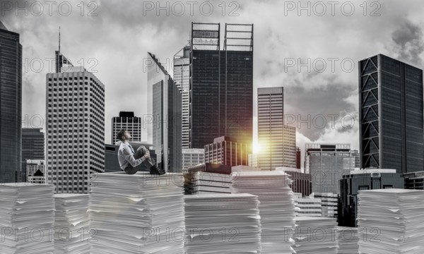 Thoughtful businessman sitting on pile of documents and looking away with cityscape and sunlight on background. Mixed media