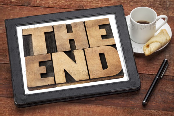 The end - text in vintage letterpress wood type on a digital tablet with a cup of coffee