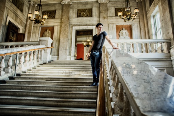 Young Handsome Man Standing on Stairs and Leaning Against Rail Inside an Architectural Building While Looking Into the Distance