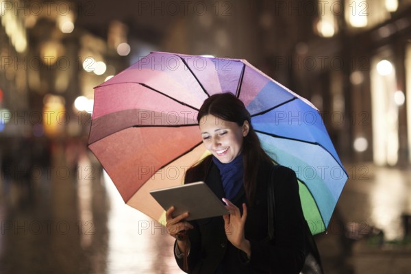 Happy young girl with colorful umbrella using tablet computer outdoor. She standing in the city street in the rainy evening