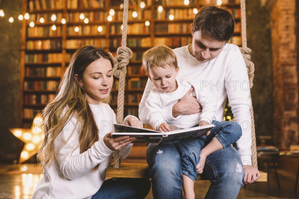 Family togetherness. Mom, dad and son reading story book together sitting on swing evening at home. Family and Parenthood Concept. Happy family reads childrens book while sitting home in loft interior