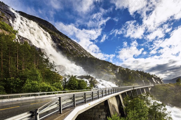 Huge powerful and roaring cascading Langfossen waterfall on the Vaule river. The bridge is fenced with railings. Passes through the highway. Summer cold and rainy day. Western Norway