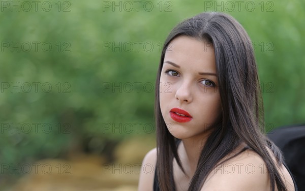 Young beautiful serious brunette girl with red lips and long straight hair over green nature background