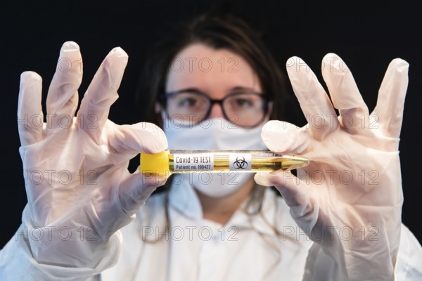 Selective focus shot of vial filled with urine sample and labeled Covid-19 test, held center frame by masked out of focus doctor over black background