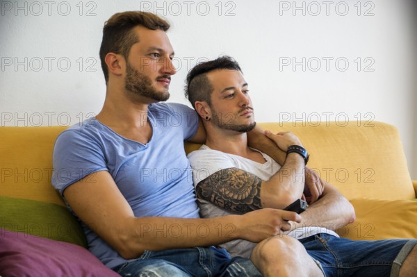Two gay men in casual outfits sitting on sofa embracing and watching TV at home