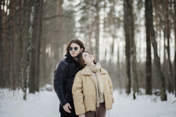 Couple in a snowy forest with the woman leaning on the man, both in winter attire among towering trees