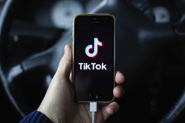 A hand holding a smartphone with TikTok app in front of a steering wheel inside a car
