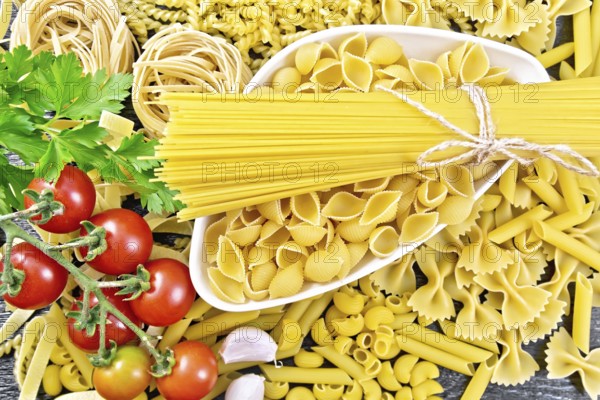 Pasta seashells and spaghetti in bowl, pipe rigate, tagliatelle, fusilli, penne, farfalle, conquilla, trenette, rotini, pappardelle, tomatoes, garlic and parsley on wooden board background from above