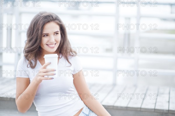 Beautiful young woman holding takeaway cup of coffee and sitting on bridge against water background