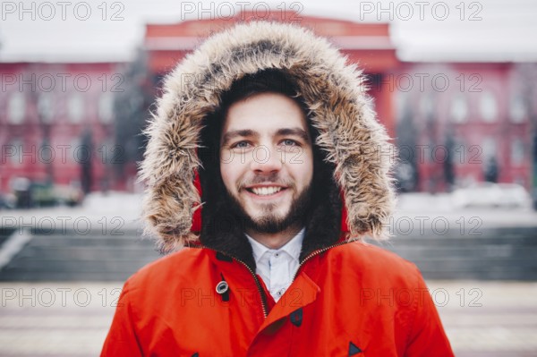 Portrait, close-up of young stylishly man smiling with a beard dressed in red winter jacket with a hood and fur on his head stands against the background of a snow-covered city. Winter and frost theme