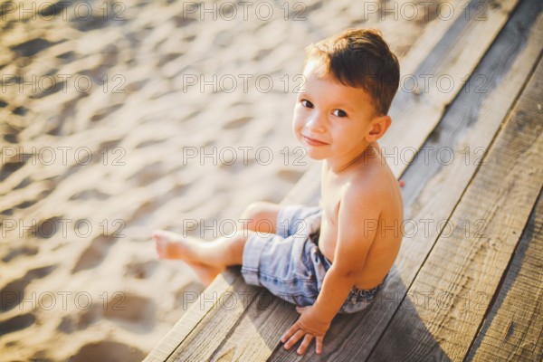 Little Caucasian boy child sitting on wooden pier sandy beach, summer time, sea holiday near water. The theme is the flow of time, a short life, the meaning and purpose of existence. Meaning of life