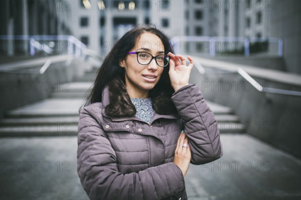 Portrait beautiful young business woman brunette in jacket and jumper stands on background office building, business centre in glasses for view. Hand holding onto shackle of glasses and looking away