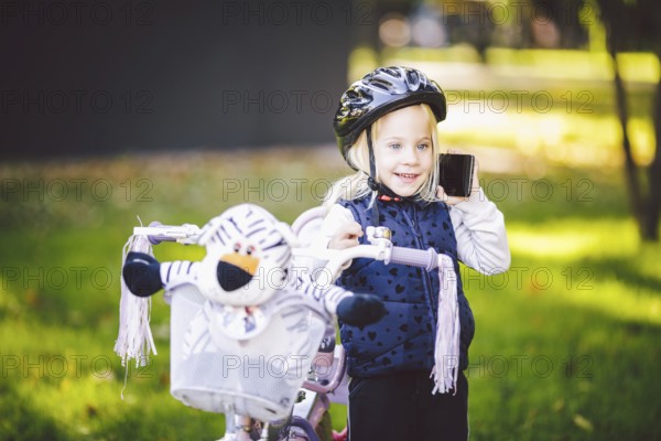 Funny child Caucasian girl blonde in a bicycle helmet near a purple bike with a basket in outside the park on a green lawn graß cart at home. Plays uses hand phone with toothy smile