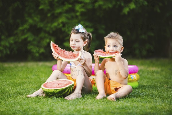 Funny little kids brother and sister eating watermelon on green graß near inflatable pool in yard at home. Toddler boy and girl. Children eating fruit in garden. Childhood, Family, Healthy Diet