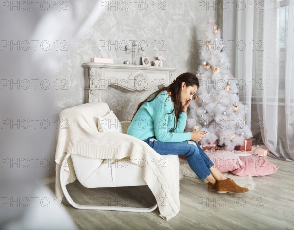 Pretty brunette woman sitting on the armchair texting on the phone near the Christmas tree