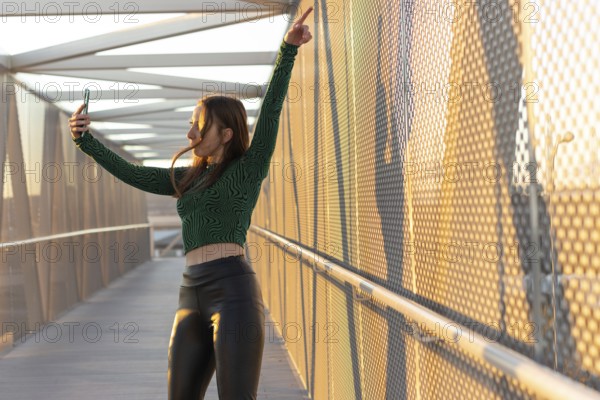 Happy young woman dancing and filming herself with her cell phone in the street at sunset.