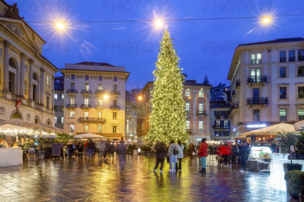 Christmas tree in Lugano, the center of the Reformation Square (Piazza della Riforma) . The disco starts (some people dance a little. Outdoor cafe and blurred motion of people. Reflections on the stone floor of the square