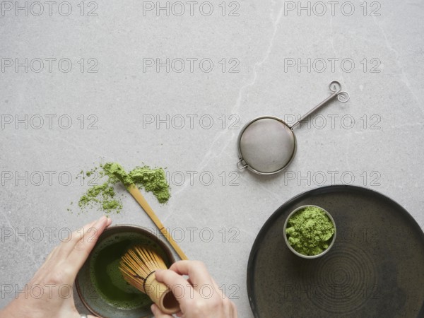 Preparation of green tea drink with matcha powder. Hands with traditional bamboo whisk over bowl of matcha tea drink on gray stone table