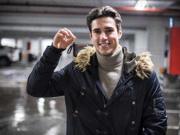 Young trendy man in winter outerwear standing on parking showing alarm remote control to the camera, smiling