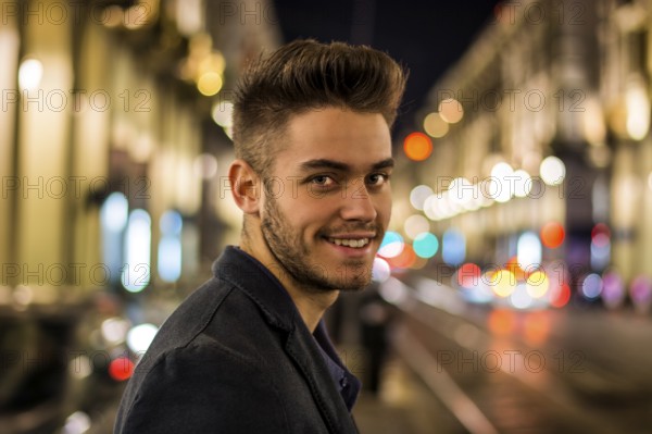 Attractive young man portrait at night with city lights behind him, smiling and looking at camera in Turin, Italy, Europe