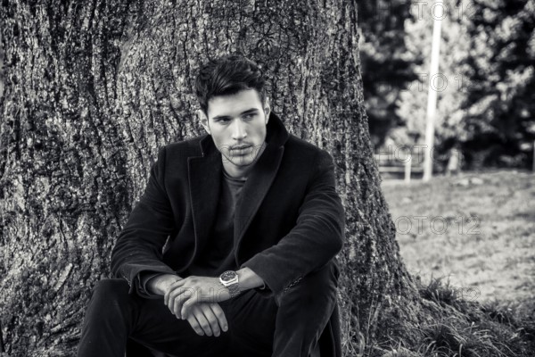 Handsome young man leaning against tree, looking to a side, in a sunny day wearing a black coat