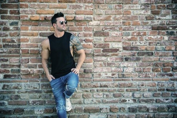 Attractive muscular man standing outdoor leaning against old brick wall, looking to a side