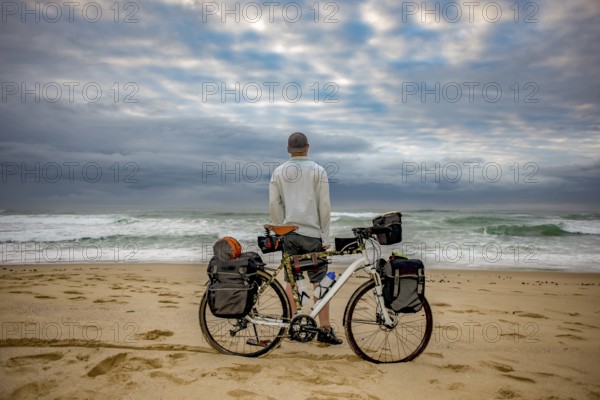 A cycle packer long distance cyclist stand in front of his bicycle on the beach while looking at the ocean