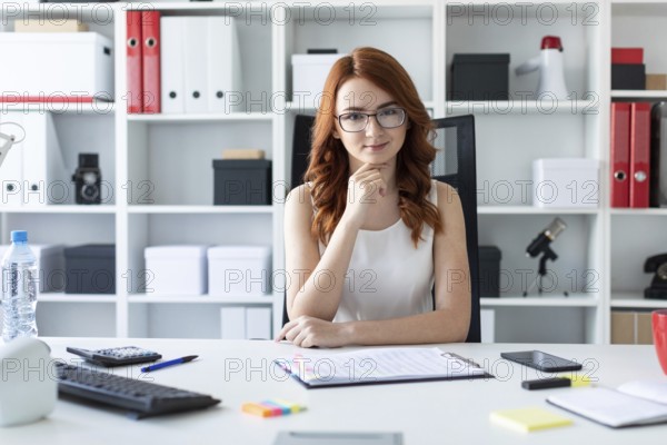A red-haired young girl with glasses, a white blouse and a red skirt is working in the office. photo with depth of field