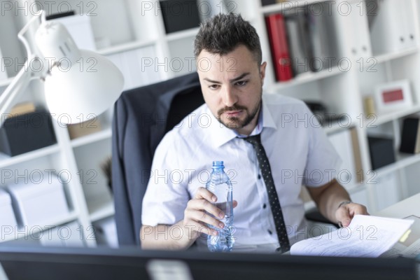 A man is sitting at a table in the office, working with documents and holding a bottle of water in his hand