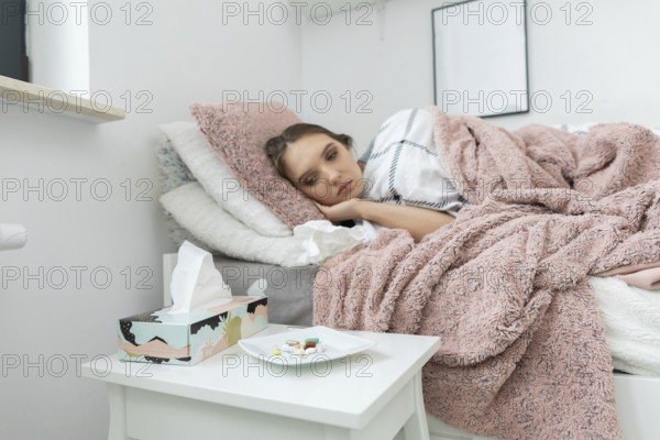 A sick teenager is lying on the bed. In front of the bed is a white table with a plate full of various tablets