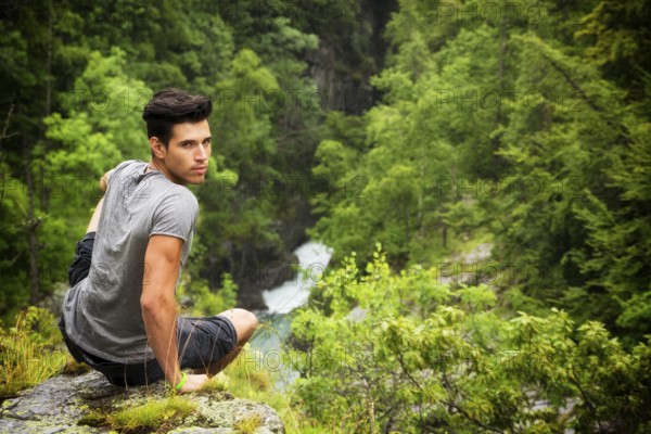 Handsome muscular young man sitting in lush green mountain scenery looking at camera