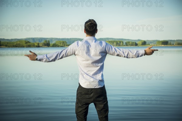 Young man in shirt, outdoor with arms spread open enjoying freedom in front of lake, seen from the back