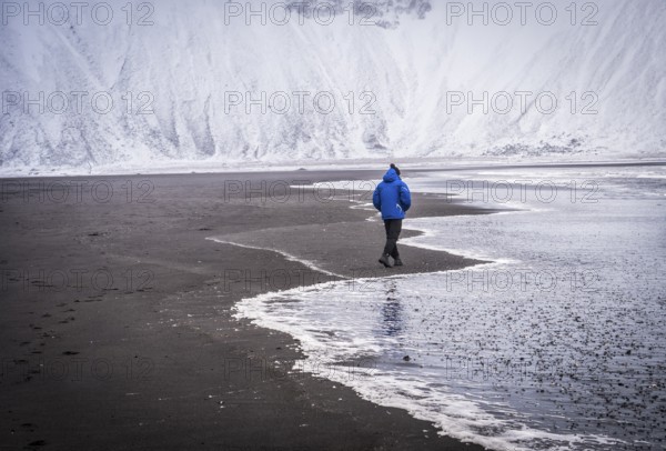 Full body back view of unrecognizable male explorer in blue warm jacket walking along waving sea near snowy mountains in Iceland