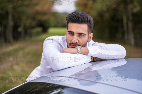 Portrait of young attractive man in business suit leaning on his new stylish car outdoor in countryside