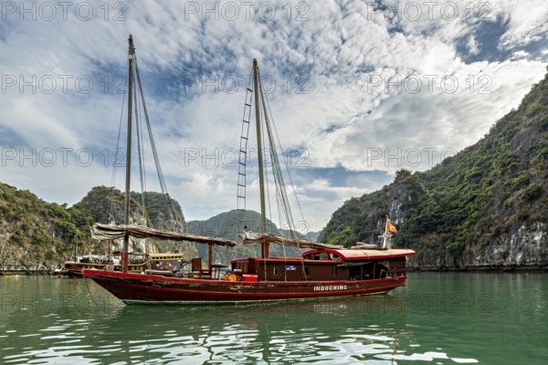 Traditional wooden boat sails on turquoise water between green hills under a cloudy sky, sailing boat for tourists in Halong Bay in Vietnam