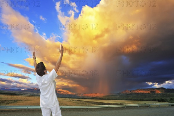 Giant cloud illuminated by the sunset. Yoga on the road. Elderly woman in white performs asana Sun salutation on the highway