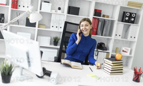 A young girl is sitting at a computer desk, holding a yellow marker in her hand and talking on the phone. Before the girl lies an open book