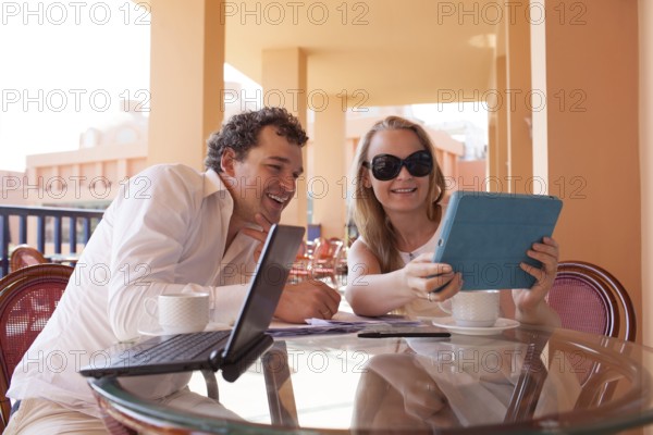Young couple relaxing over coffee on a balcony sitting at a glass table smiling as they lean close together to read the screen on a tablet computer