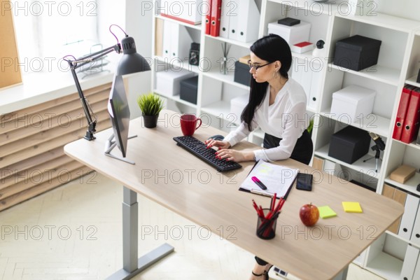 A young girl with glasses, wearing a white blouse and black trousers, works in the office. The girl has long straight black hair. photo with depth of field