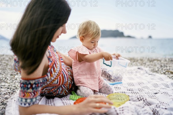 Mom lies on a blanket on a pebble beach next to a sitting little girl.