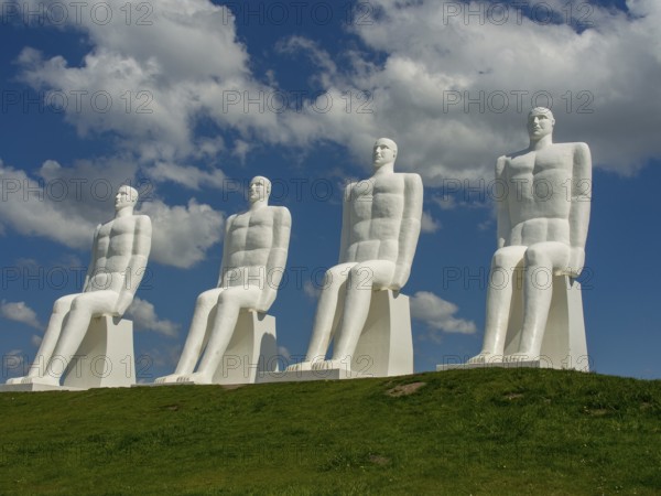 Four large white sculptures of seated men on a green meadow under a blue sky with clouds, esbjerg, denmark