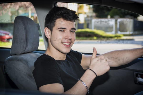 Happy young man or teenager doing thumb up sign while driving car and smiling at camera