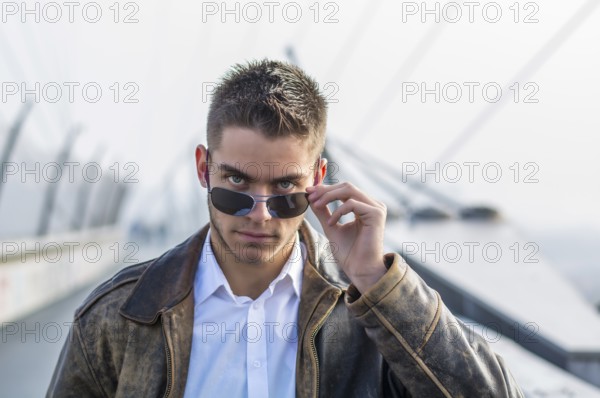 Handsome trendy young man standing on a sidewalk, wearing sunglasses and a fashionable leather jacket, in a relaxed confident pose looking at camera over the glasses