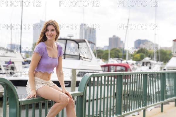 A gorgeous redhead model enjoys the perfect summer weather at a local marina, surrounded by boats of all kinds in the background, their reflections dancing on the water under the bright, sunny sky, Milwaukee, United States of America